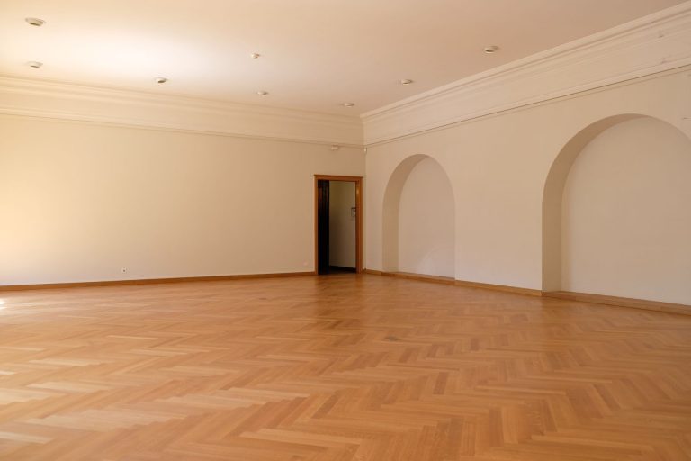 A large empty room with elegant wooden flooring, arches, and natural lighting